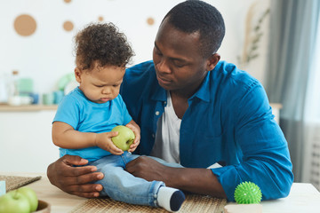 Portrait of happy African-American father playing with cute little boy on table with green apples in home interior, copy space