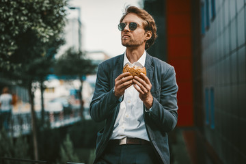 Handsome trainee having a lunch break near his office. Young man eats hamburger outside of his business center. Non healthy food concept.