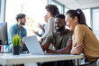 © Sanja - A man and a woman laughing while looking at the lap top with another man and a woman in the background of a workspace environment.