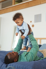 Wall Mural - Vertical portrait of happy African-American dad playing with baby son while laying on bed at home