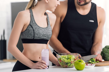 cropped view of cheerful woman holding glass of smoothie near man cooking in kitchen