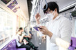 © zephyr_p - Asian man wearing surgical face mask using smartphone on skytrain or urban train. Wuhan coronavirus (COVID-19) outbreak prevention in public transportation. Health awareness for pandemic protection