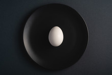 Single White Egg In Black Plate On Dark Moody Plain Minimal Background, Top View, Happy Easter Day
