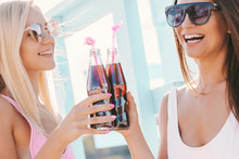 Portrait Of Two Happy Girls Friends Drinking Cold Refreshing Lemonade And Laughing At Sunny Beach. Beautiful Cheerful Women In Stylish Swimsuits And Sunglasses Cheering With Bottles Of Soft Drink