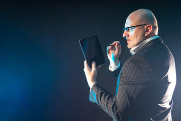 Wall Mural - Human with a tablet in his hands. A man in a business suit and glasses. Adult businessman on a dark background. Concept - A man works on a tablet. The gadget is off. Modern businessman