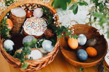 Easter modern eggs, easter bread, ham, beets, butter, in wicker basket decorated with green buxus branches and flowers on rustic wooden table. Traditional Easter basket.