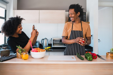 Wall Mural - Afro woman taking photo of her husband while he prepares dinner.