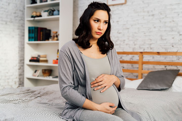 Asian mixed Caucasian concerned and anxious woman in pregnancy overthinking sits on bed at home. Emotional and insecure after pregnancy problem concept