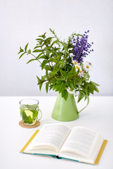 eco and organic concept - cup of green herbal tea, book and flowers in jug on table