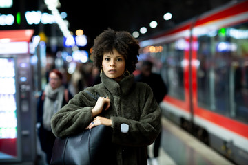 Stylish portrait of a beautiful African-American woman with black curly hair in a fur coat. Woman standing on urban city subway metro platform at night. Fashion and lifestyle