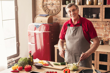 Wall Mural - Smiling Aged Man Preparing Fresh Vegetable Salad, Enjoying Cooking In Kitchen