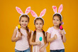 © producer - happy cute little child girls with pink bunny ears holding painted Easter eggs on studio yellow background. Easter day