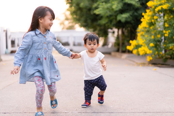 Wall Mural - Adorable asian little toddler boy is holding hand with his big sister while he try to practice for first step, concept of love and relation of sibling in family, trust, friendship and outdoor activity