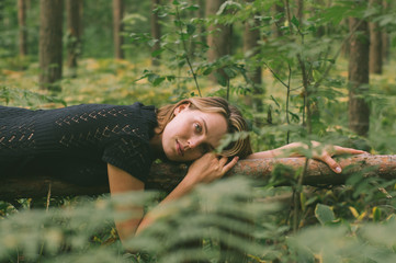 Portrait of young beautiful girl in black dress in forest that lies on the tree trunk