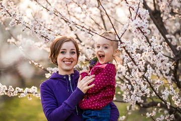 Young mother and child daughter together, hugging and laughing in a Flowering apricot garden. Family outdoors lifestyle.