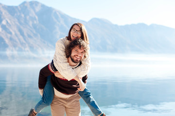 Happy couple in love hugging at winter sea beach. Travelers smiling by blue mountains and lake. Young man giving woman piggyback in vacation, outdoor adventure. Romantic lifestyle moment. Copy space