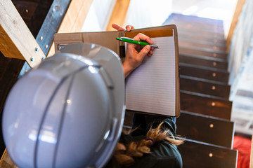 top view of construction inspector woman sits on the stairs during a home pre-purchase inspection, she uses clipboard to writes notes. with copy space