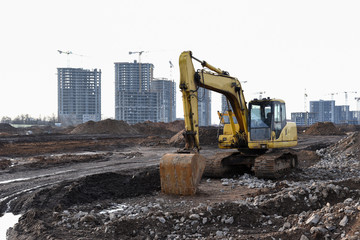 Wall Mural - Yellow excavator with bucket at building under construction. Backhoe at large scale construction site against tower cranes.  Digging the ground for the foundation and for laying sewer pipes