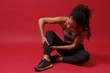 Worried young african american sports fitness woman in sportswear posing working out isolated on red background studio portrait. Sport exercises healthy lifestyle concept. Sitting, touching ankle.