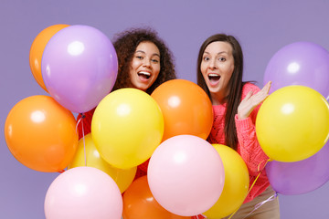 Excited two european african american women friends in knitted sweaters isolated on violet purple background. Birthday holiday party, people emotions concept. Celebrating hold colorful air balloons.