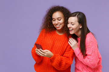 Sticker - Smiling two young european african american women friends in knitted sweaters isolated on violet purple background. People sincere emotions lifestyle concept. Using mobile phone, typing sms message.