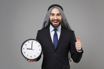 Wall Mural - Cheerful young bearded arabian muslim businessman in keffiyeh kafiya ring igal agal classic suit isolated on gray background. Achievement career wealth business concept. Hold clock showing thumb up.