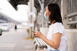 © DG PhotoStock - Asian young woman waiting for the bus at bus stop in the crowded and busy city and touching on smartphone for searching a map. City where is fully with air pollution and crowd. Environment problem.