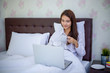 © tuiphotoengineer - Asian woman is drinking a cup of coffee and using a notebook to work on the bed.She smiles and enjoys working at home.