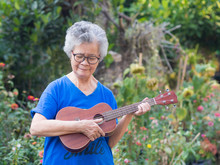 Portrait Of Elderly Woman Playing Ukulele In Her Garden. Relaxing By Singing And Play Small Guitar Happy And Enjoy Life After Retired. Concept Of Old People And Health Care. Space For Text