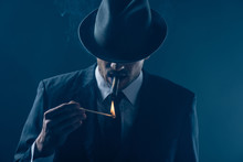 Mafioso With Covered Eyes With Felt Hat Lighting Cigar On Dark Blue Background