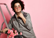 Girl In Sunglasses, Gray Shirt Dress And Pants. Smiling, Standing On Metal Ladder, Holding Bouquet, Posing On Pink Background