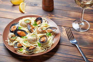 Seafood on a brown plate. Mussels, snails and pasta with parsley. Nearby is a glass of wine, white bread, lemon and a fork. Close-up