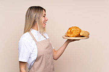 Wall Mural - Young blonde girl with apron. Female baker holding a table with several breads with happy expression