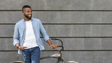 Handsome Black Man Standing With Bike Against Urban Wall, Looking Away