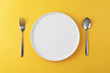Empty white plate with spoon and fork on yellow background