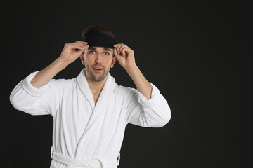 Wall Mural - Surprised young man in bathrobe and eye sleeping mask on black background. Space for text