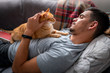 gato atigrado se encuentra en el pecho de un joven tumbado en el sofá debajo de la ventana