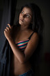 © abir - Portrait of young and beautiful dark skinned Indian Bengali woman in colorful lingerie/bikini and hot pants standing in front of window and looking outside in studio background. Boudoir photography.