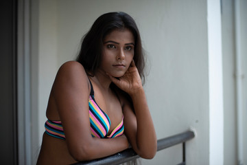 Portrait of an young and beautiful dark skinned Indian Bengali woman in colorful lingerie/bikini and hot pants posing in casual mood on a balcony in white urban background. Boudoir photography.