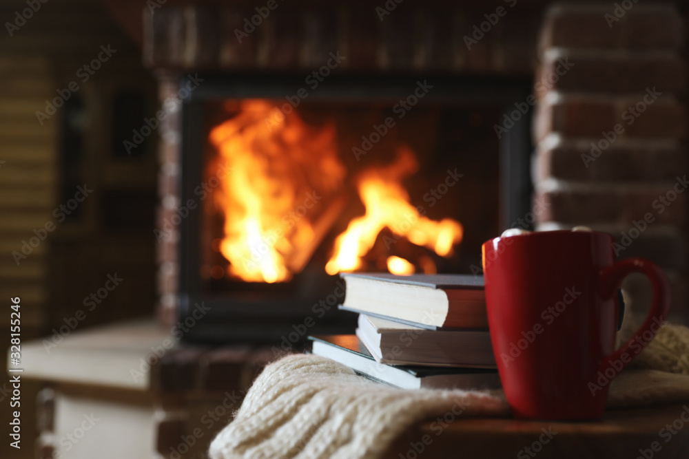 Books and cocoa on table near burning fireplace, space for text. Reading at home - obrazy, fototapety, plakaty 
