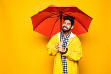 Young Handsome Bearded Indian Man In Yellow Raincoat With Red Umbrella Cover From Rain Isolated Over Orange Background