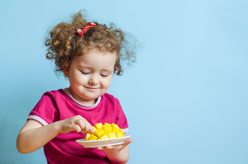 little curly girl with an appetite eats mango on a blue background with copy space. baby, toddler ta