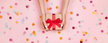Banner. Woman Hands Holding Present Box With Red Bow On Pastel Pink Background With Multicolored Confetti. Flat Lay Style.