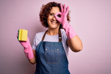 Poster - Middle age curly hair woman cleaning doing housework wearing apron and gloves using scourer with happy face smiling doing ok sign with hand on eye looking through fingers