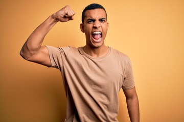 Young handsome african american man wearing casual t-shirt standing over yellow background angry and mad raising fist frustrated and furious while shouting with anger. Rage and aggressive concept.