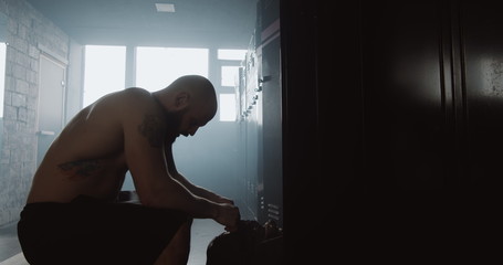 Exhausted young shirtless man sitting alone in dark gym locker room feeling sad after failure day. Good fighting spirit.