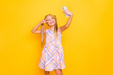 Photo of pretty small lady hold telephone young influencer making selfies for blog showing v-sign symbol near eye wear checkered summer dress isolated bright color background