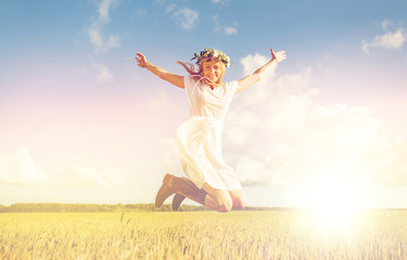 happiness, nature, summer holidays, vacation and people concept - smiling young woman in wreath of flowers and gumboots jumping on cereal field