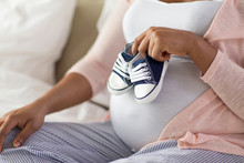 Pregnancy, People And Rest Concept - Close Up Of Pregnant African American Woman Sitting On Bed And Holding Little Baby Bootees At Home Bedroom