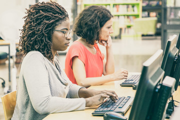 Wall Mural - Multiracial adult students working in computer class. Women in casual sitting at table, using desktops, typing, looking at monitor. Online course concept